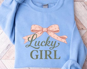 May include: A light blue sweatshirt with a peach-colored bow graphic and the words "Lucky GIRL" in a script font. The bow has a dotted pattern. The sweatshirt has long sleeves and a crew neck.