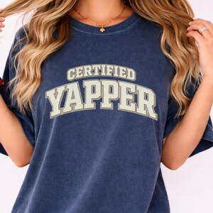 May include: Navy blue t-shirt featuring the text "CERTIFIED YAPPER" in a vintage, collegiate-style font. The shirt has short sleeves and a relaxed fit. The shirt is made of a soft, comfortable material.
