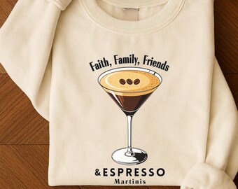 May include: Cream-colored sweatshirt featuring a graphic of an espresso martini with the words "Faith, Family, Friends" above and "& Espresso Martinis" below. The martini is in a classic glass with three coffee beans on top.