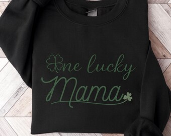 May include: Black sweatshirt featuring the green text "One lucky Mama" with a shamrock. The garment is folded, showing the sleeves. The design is centrally placed on the front of the sweatshirt.