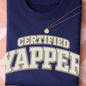 May include: A navy blue sweatshirt featuring the text "CERTIFIED YAPPER" in a retro font. A gold necklace with a circular pendant is draped across the shirt. The sweatshirt is neatly folded on a wooden surface.