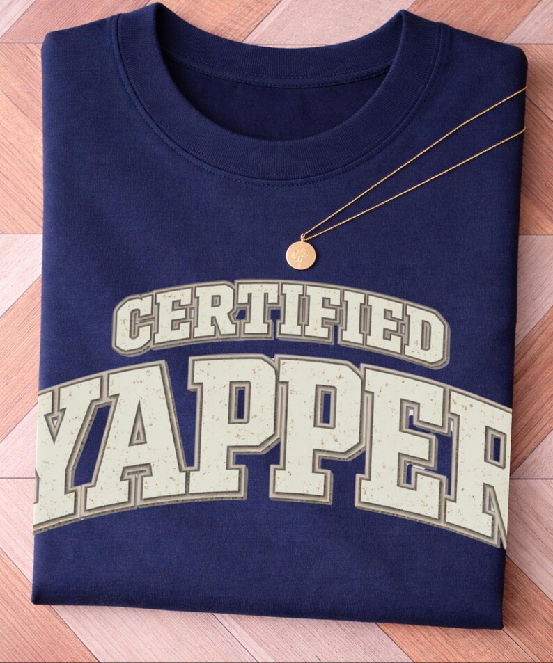 May include: A navy blue sweatshirt featuring the text "CERTIFIED YAPPER" in a retro font. A gold necklace with a circular pendant is draped across the shirt. The sweatshirt is neatly folded on a wooden surface.
