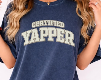 May include: Navy blue t-shirt with the text "CERTIFIED YAPPER" in a vintage-style, off-white font. The shirt has short sleeves and a crew neck. The overall style is casual and relaxed.