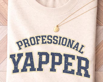 May include: A cream-colored crew neck t-shirt featuring the text "PROFESSIONAL YAPPER" in a distressed navy blue collegiate font. A thin gold chain necklace with a small round pendant rests on the upper chest area of the shirt.