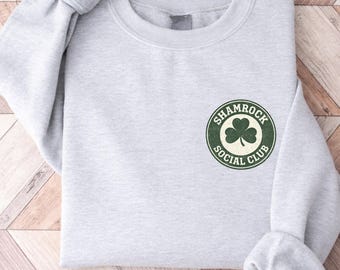 May include: A light gray sweatshirt with the text "SHAMROCK SOCIAL CLUB" in a green and white circular design. The sweatshirt is folded on a wooden surface.