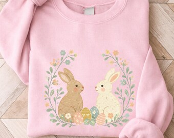 May include: A light pink sweatshirt featuring a floral wreath design with two bunnies and Easter eggs. The bunnies are light brown and cream-colored. The wreath includes green leaves and small yellow and blue flowers.