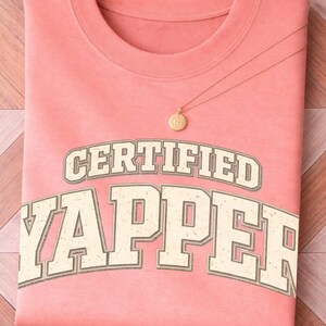 May include: A folded coral t-shirt with the text "CERTIFIED YAPPER" in a vintage font. A gold necklace with a circular pendant rests on the shirt. The shirt is on a wooden surface.