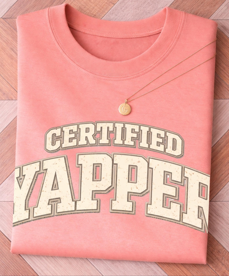May include: A folded coral t-shirt with the text "CERTIFIED YAPPER" in a vintage font. A gold necklace with a circular pendant rests on the shirt. The shirt is on a wooden surface.