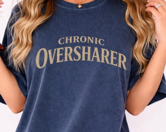May include: Navy blue t-shirt with the words "CHRONIC OVERSHARER" printed in a tan color. The shirt has short sleeves and a crew neck. The fabric appears to be soft and slightly faded.