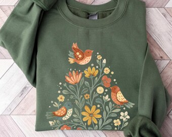 May include: An olive green sweatshirt featuring a floral and bird design. The graphic includes colorful flowers and three birds in shades of orange, yellow, and blue. The sweatshirt has long sleeves with gathered cuffs.