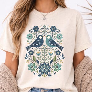 May include: Cream-colored t-shirt featuring a floral design with two birds facing each other. The birds are blue and teal, surrounded by blue, teal, and yellow flowers and leaves. The shirt has a crew neck and short sleeves.