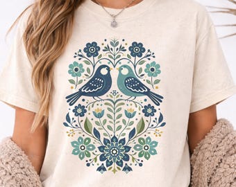 May include: Cream-colored t-shirt featuring a floral design with two birds facing each other. The birds are blue and teal, surrounded by blue, teal, and yellow flowers and leaves. The shirt has a crew neck and short sleeves.