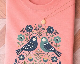 May include: A coral-colored t-shirt with a floral and bird design. Two birds face each other amidst blue flowers and green leaves. A gold necklace with a circular pendant rests on the shirt.