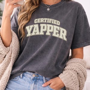 May include: A dark grey t-shirt featuring the text "CERTIFIED YAPPER" in a cream-coloured, collegiate-style font. The shirt is paired with blue jeans and a cream-coloured cardigan.