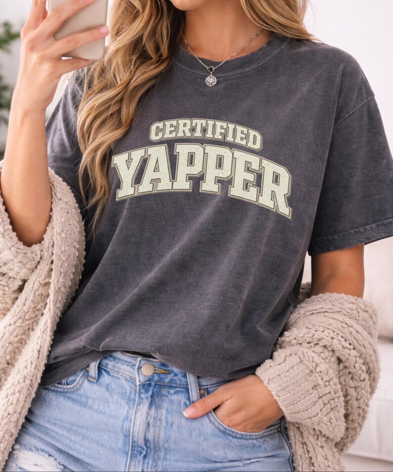 May include: A dark grey t-shirt featuring the text "CERTIFIED YAPPER" in a cream-coloured, collegiate-style font. The shirt is paired with blue jeans and a cream-coloured cardigan.