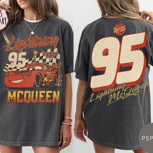 May include: A charcoal gray t-shirt featuring a graphic of the red race car Lightning McQueen with the number 95, a checkered flag, and the text Lightning McQueen on the front, and a large number 95 with Rust-eze branding on the back.