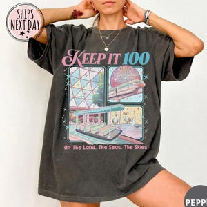 May include: Dark gray t-shirt with a colorful graphic design. The design includes the text "KEEP IT 100" and illustrations of a monorail, a boat, and a geodesic sphere. The phrase "On The Land, The Seas, The Skies" is also included.