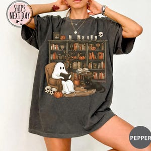 May include: A charcoal gray t-shirt featuring a graphic print of a white ghost sitting in a brown armchair reading a book. The scene includes a bookshelf filled with books, pumpkins, candles, a black cat, and skulls on a wooden floor.