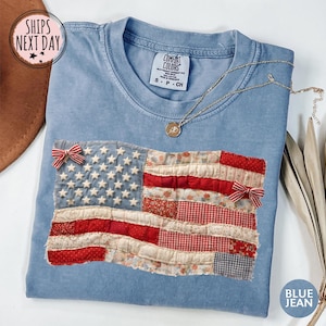 May include: A light blue t-shirt with a sewn-on American flag design. The flag features white stars on a blue field and red and white stripes. Two small red and white bows are attached to the flag. A gold necklace with a pendant is on the shirt.