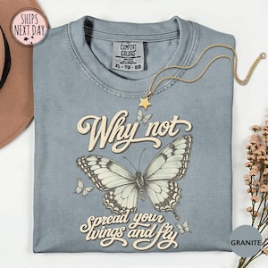 May include: A granite-colored t-shirt featuring a large central butterfly illustration with smaller butterflies surrounding it. The text reads Why not spread your wings and fly in a cream-colored script font. The shirt has a crew neckline.