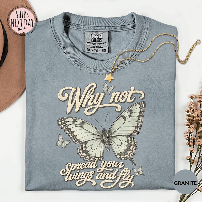 May include: A granite-colored t-shirt featuring a large central butterfly illustration with smaller butterflies surrounding it. The text reads Why not spread your wings and fly in a cream-colored script font. The shirt has a crew neckline.