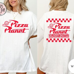May include: White t-shirt featuring the red "Pizza Planet" logo on the front and back. The back of the shirt also displays the text "DELIVERY SHUTTLE" and "SERVING YOUR LOCAL STAR CLUSTER" in red, with a red and white chequered pattern.