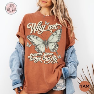 May include: A rust-colored short-sleeve t-shirt featuring a large central butterfly graphic, smaller butterflies, and the text Why not spread your wings and fly in a cream-colored script font. The shirt is worn with a light blue denim jacket.