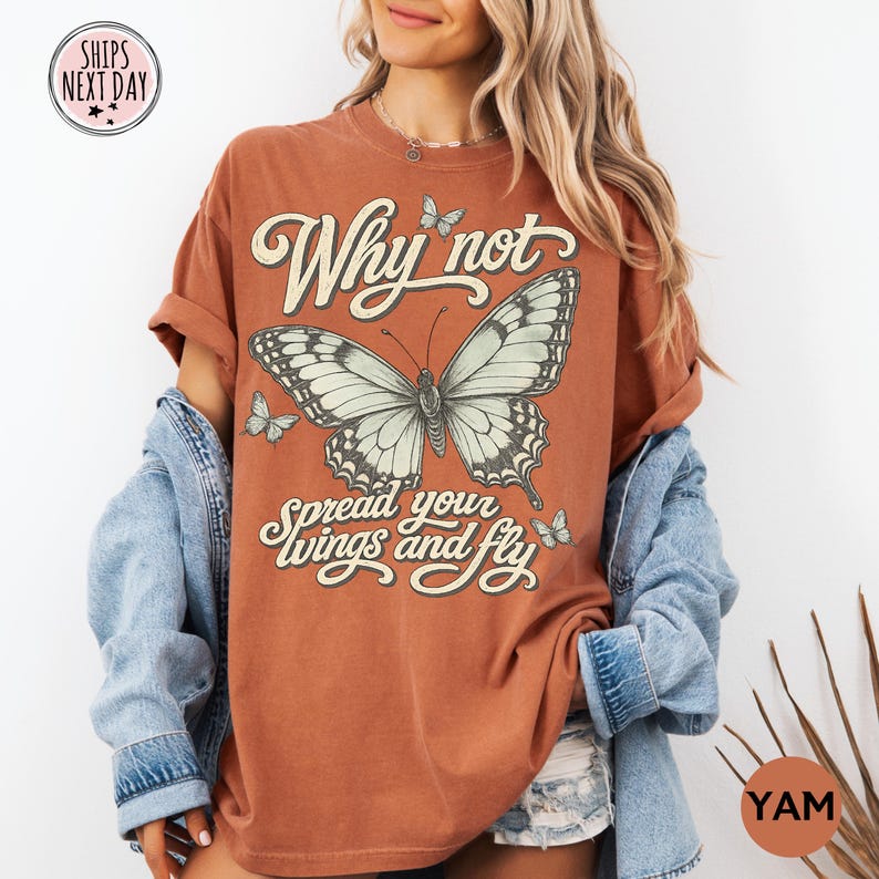 May include: A rust-colored short-sleeve t-shirt featuring a large central butterfly graphic, smaller butterflies, and the text Why not spread your wings and fly in a cream-colored script font. The shirt is worn with a light blue denim jacket.