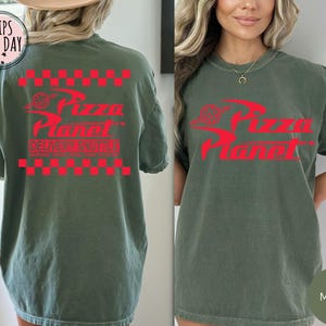 May include: A moss green t-shirt featuring a red Pizza Planet logo and text. The design includes a red checkered pattern above and below the logo, with the words "Delivery Shuttle" below. The shirt has a relaxed, oversized fit.