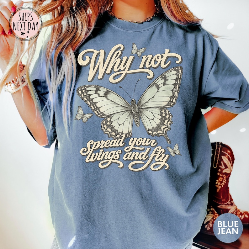 May include: A blue t-shirt featuring a large central butterfly graphic with smaller butterflies surrounding it. The text "Why not spread your wings and fly" is printed in a cream-colored script font. The shirt has short sleeves and a crew neckline.