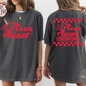 May include: Dark grey t-shirt featuring a red "Pizza Planet" logo on the front and back. The back of the shirt also has the text "Delivery Shuttle" and a red and white chequered pattern. The shirt has a relaxed fit.