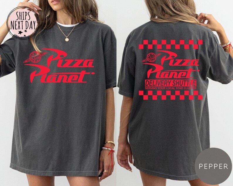 May include: Dark grey t-shirt featuring a red "Pizza Planet" logo on the front and back. The back of the shirt also has the text "Delivery Shuttle" and a red and white chequered pattern. The shirt has a relaxed fit.