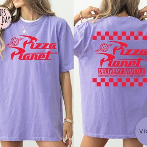 May include: A violet t-shirt featuring the red "Pizza Planet" logo on the front and back. The back also displays "Delivery Shuttle" text and a red and white checkered pattern. A casual, unisex t-shirt.