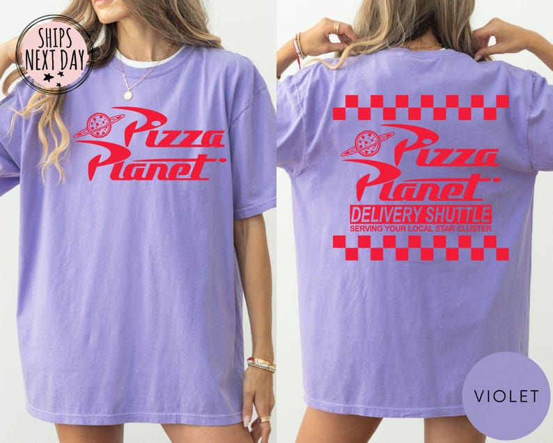 May include: A violet t-shirt featuring the red "Pizza Planet" logo on the front and back. The back also displays "Delivery Shuttle" text and a red and white checkered pattern. A casual, unisex t-shirt.