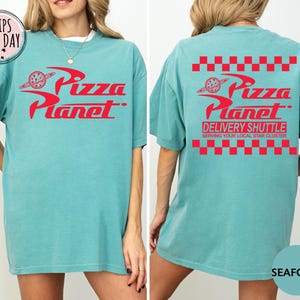 May include: Seafoam green t-shirt with the red "Pizza Planet" logo on the front and back. The back also features "Delivery Shuttle" text and a red checkered pattern. A fun, casual tee.