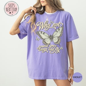 May include: A light purple short-sleeve t-shirt featuring a large central butterfly graphic with the text Why not spread your wings and fly in a decorative font. The design includes smaller butterflies surrounding the main illustration.