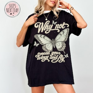 May include: A black short-sleeve t-shirt featuring a large central butterfly graphic with the text Why not spread your wings and fly in a cream-colored script font. Smaller butterfly illustrations are positioned above and below the text.