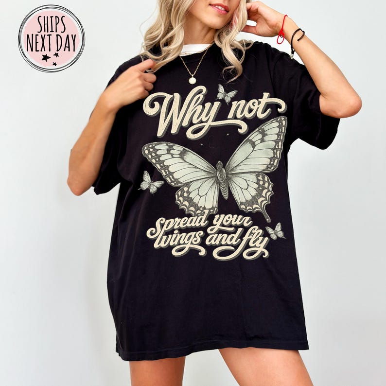May include: A black short-sleeve t-shirt featuring a large central butterfly graphic with the text Why not spread your wings and fly in a cream-colored script font. Smaller butterfly illustrations are positioned above and below the text.