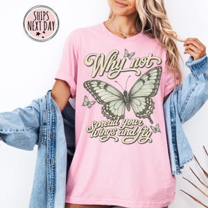 May include: A light pink short-sleeve t-shirt featuring a large central butterfly graphic with smaller butterflies surrounding it. The text reads Why not spread your wings and fly in a decorative font. The shirt is worn with a blue denim jacket.