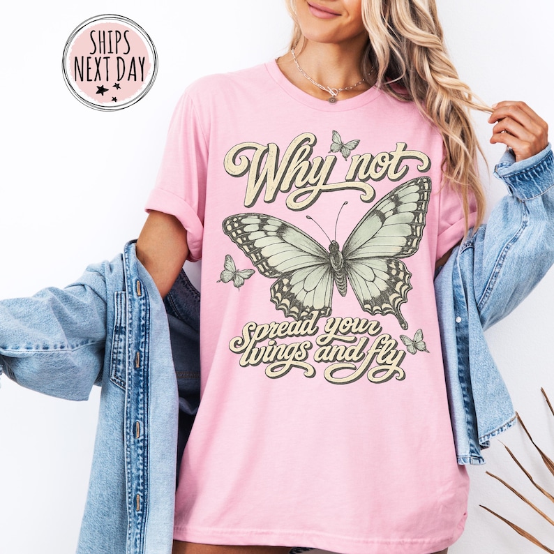May include: A light pink short-sleeve t-shirt featuring a large central butterfly graphic with smaller butterflies surrounding it. The text reads Why not spread your wings and fly in a decorative font. The shirt is worn with a blue denim jacket.