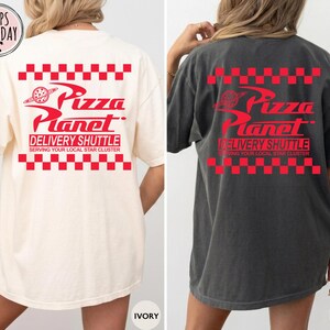 May include: Ivory and pepper-coloured t-shirts with the Pizza Planet logo in red. The design includes the text "Pizza Planet Delivery Shuttle" and "Serving Your Local Star Cluster" with a red checkered pattern.