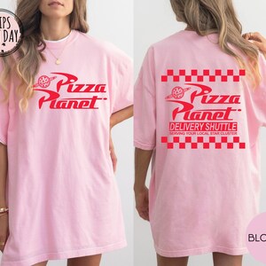 May include: A pale pink t-shirt featuring the Pizza Planet logo in red on the front and back. The back of the shirt also displays the text "DELIVERY SHUTTLE" and "SERVING YOUR LOCAL STAR CLUSTER". The shirt has a white collar.