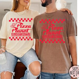 May include: Two short-sleeved t-shirts, one cream and one brown, display the Pizza Planet logo in red. The logo includes the text "Pizza Planet Delivery Shuttle Serving Your Local Star Cluster" in a red checkered pattern.