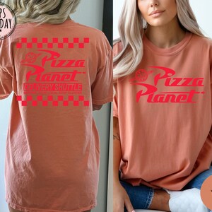 May include: A coral-coloured t-shirt featuring the Pizza Planet logo in red. The design includes the words "Pizza Planet" and "Delivery Shuttle" with a checkered pattern. The shirt is a relaxed fit.