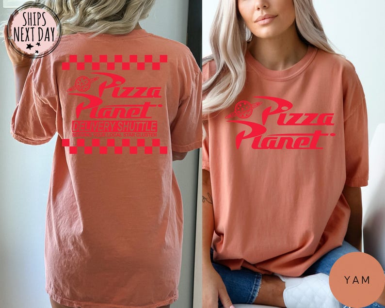 May include: A coral-coloured t-shirt featuring the Pizza Planet logo in red. The design includes the words "Pizza Planet" and "Delivery Shuttle" with a checkered pattern. The shirt is a relaxed fit.