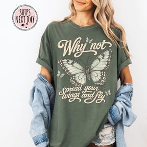 May include: An olive green short-sleeve t-shirt featuring a large central butterfly graphic with smaller butterflies surrounding it. The text "Why not spread your wings and fly" is printed in a cream-colored script font across the chest area.