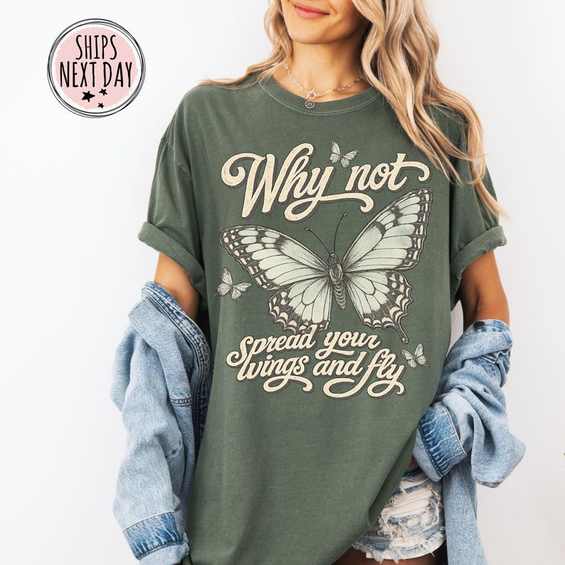 May include: An olive green short-sleeve t-shirt featuring a large central butterfly graphic with smaller butterflies surrounding it. The text "Why not spread your wings and fly" is printed in a cream-colored script font across the chest area.