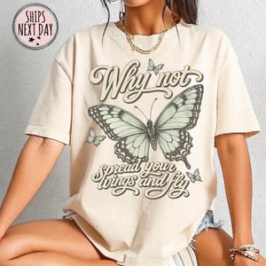 May include: A beige short-sleeve t-shirt featuring a large central butterfly graphic with the text Why not spread your wings and fly. The design includes smaller butterflies surrounding the central image in a dark brown script font.