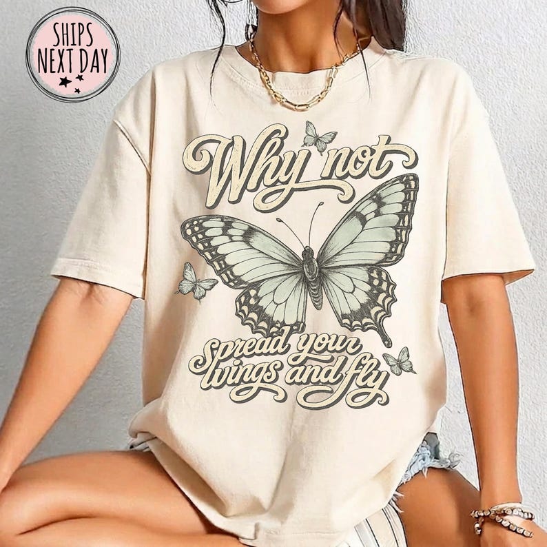 May include: A beige short-sleeve t-shirt featuring a large central butterfly graphic with the text Why not spread your wings and fly. The design includes smaller butterflies surrounding the central image in a dark brown script font.