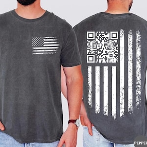 May include: Dark grey t-shirt with a distressed white American flag graphic on the front left chest. The back of the shirt features a white QR code and vertical stripes, also distressed. The shirt is a crew neck and has short sleeves.